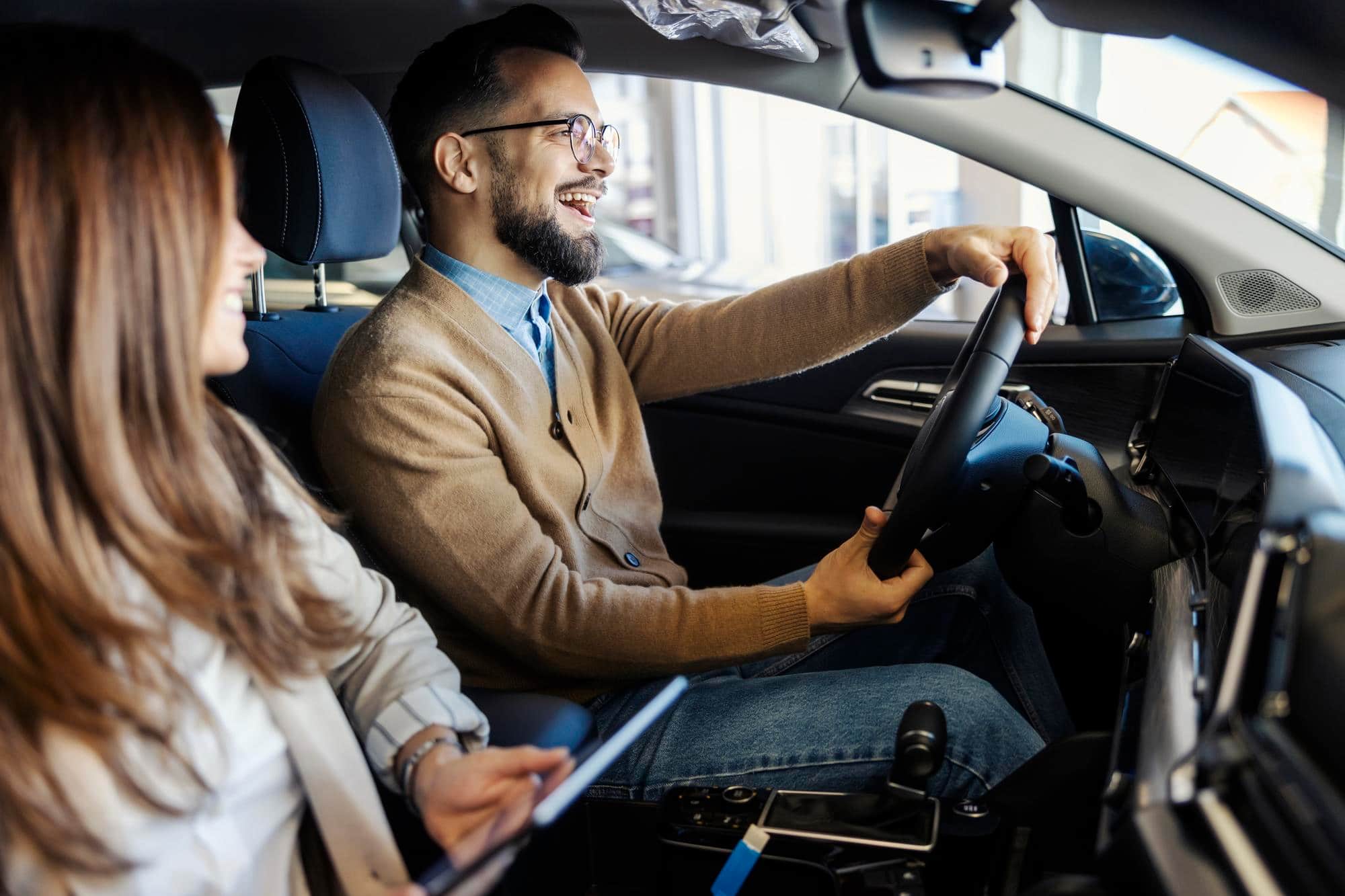 Smiling man drives car while woman in front seat holds tablet.