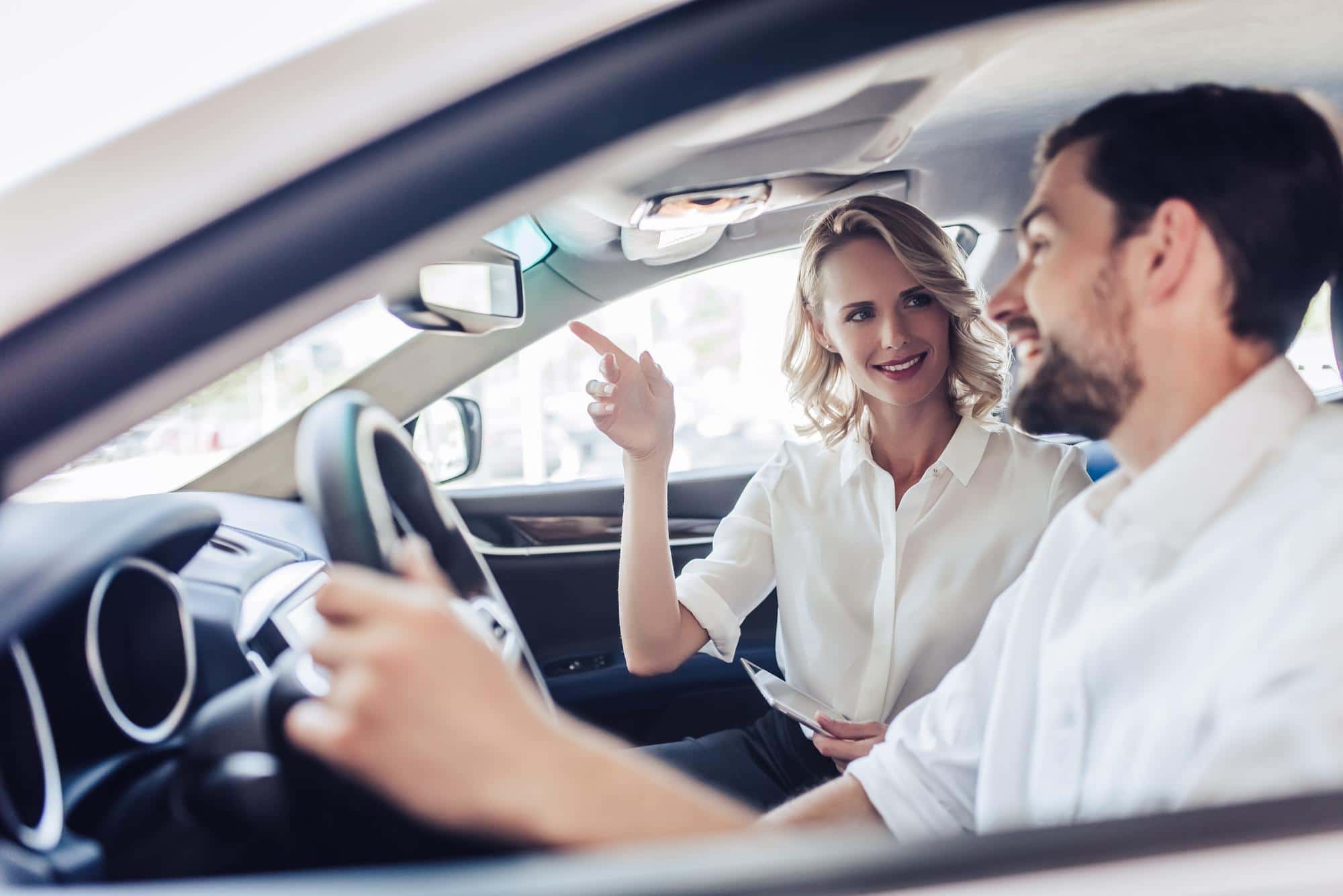 Woman in car passenger seat smiles, points at mirror, talking with happy driver.