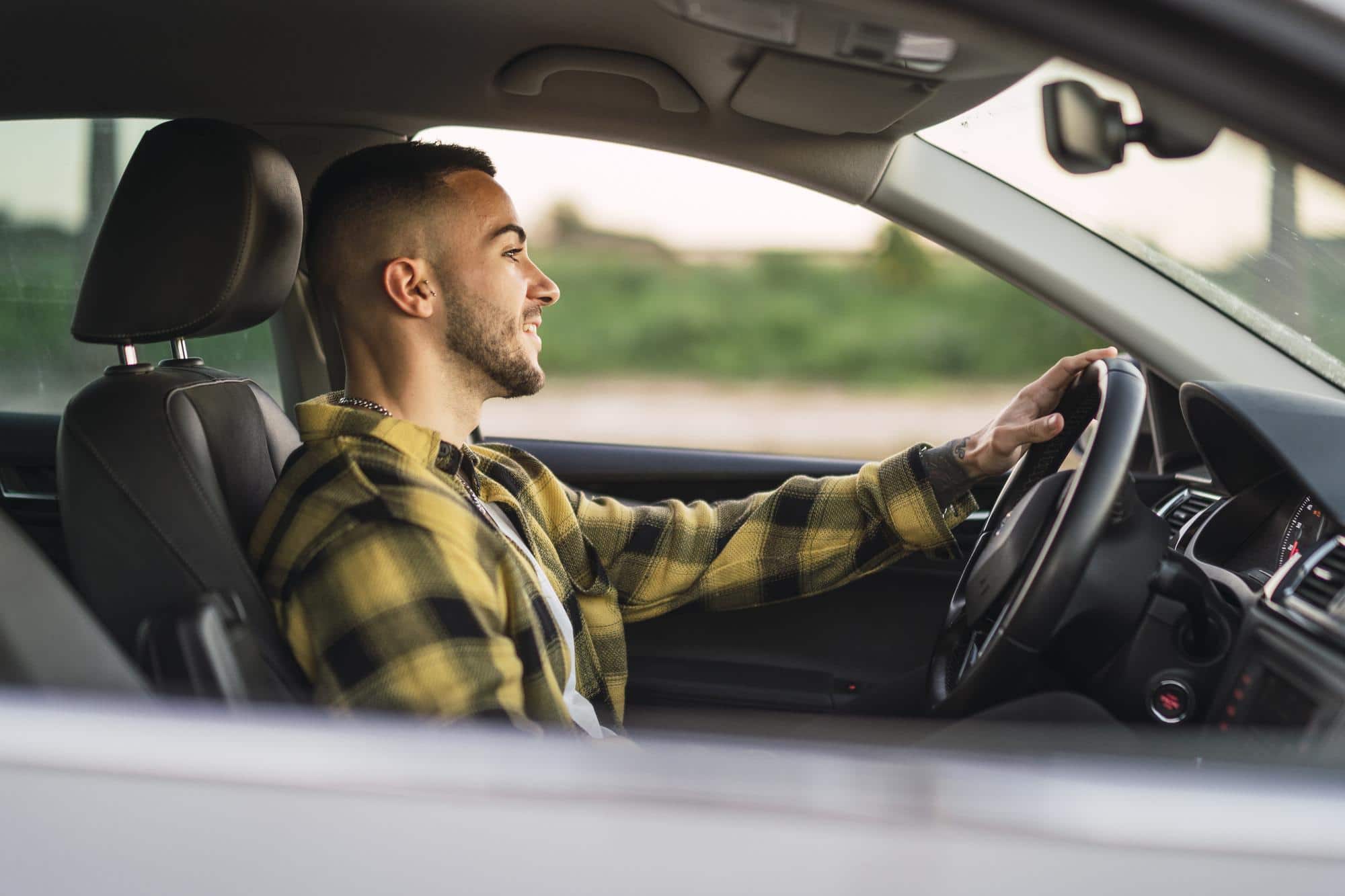 Young man in yellow plaid shirt smiling while driving, blurred scenery outside.
