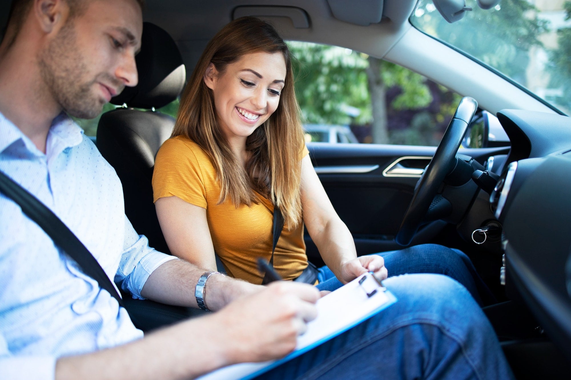 Smiling woman in driver’s seat, instructor filling out form during lesson.