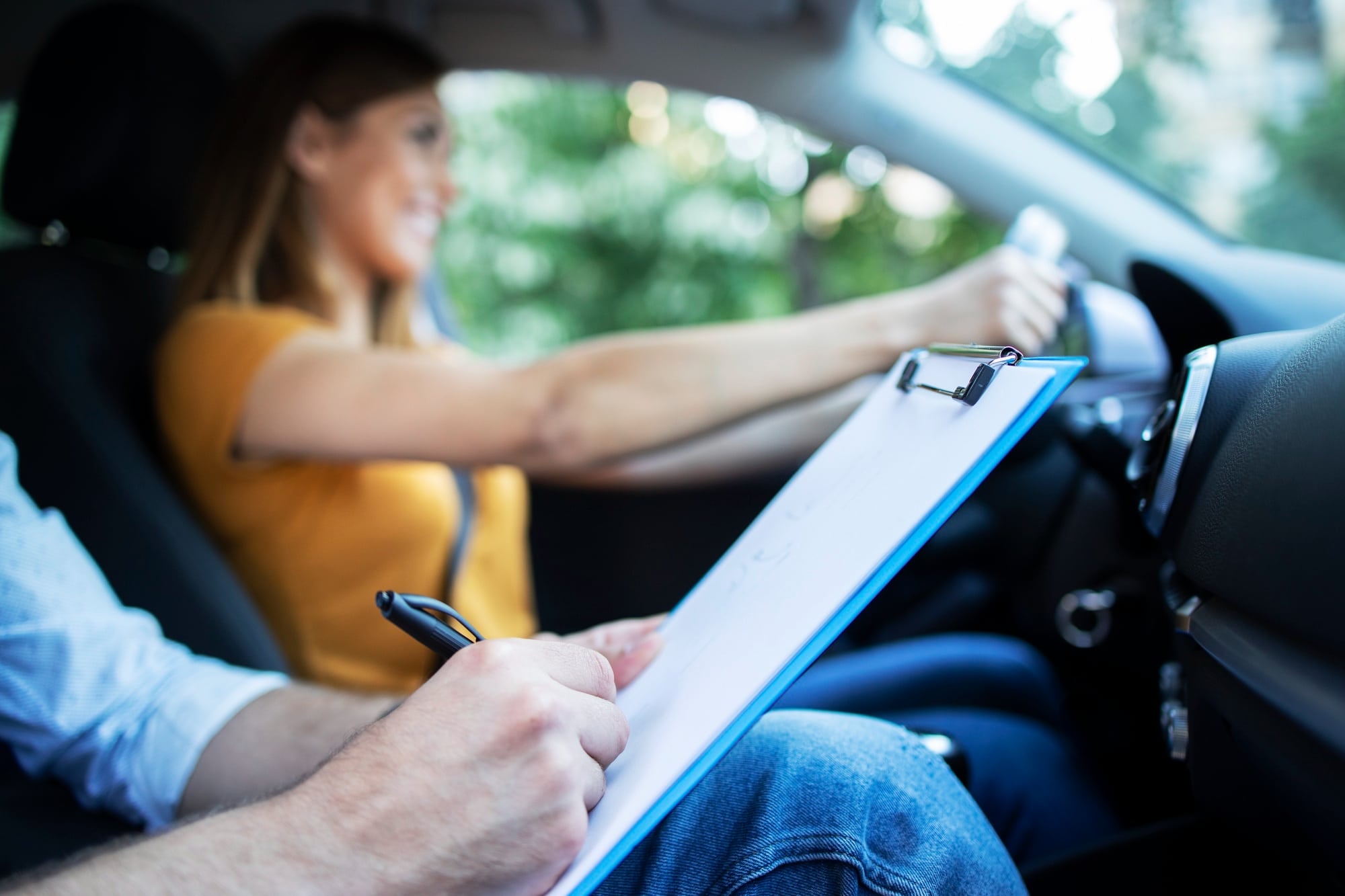 Smiling woman drives as passenger takes notes, likely during a driving lesson.
