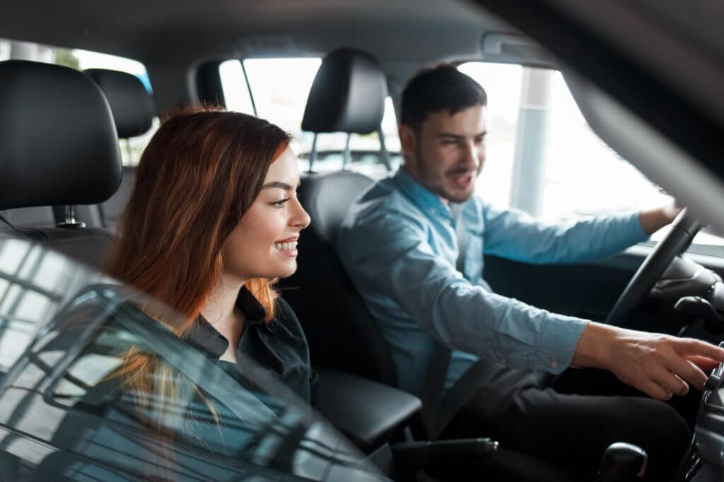 Man points at dashboard while driving; woman smiles in passenger seat. Relaxed.