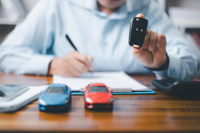 Someone writes at a desk, holding a key fob near model cars, notebook, and calculator.