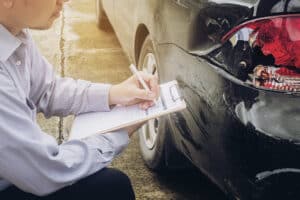 Person with clipboard inspects black car’s dented rear and broken taillight.