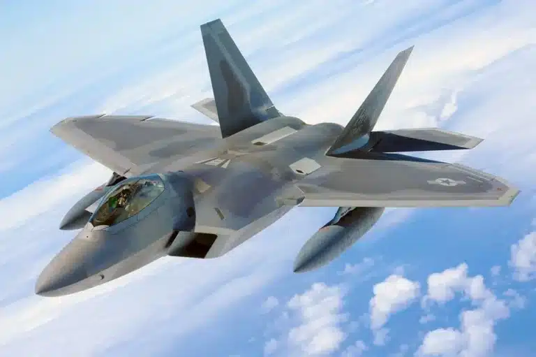 Gray fighter jet with angular design flies high above clouds in blue sky.