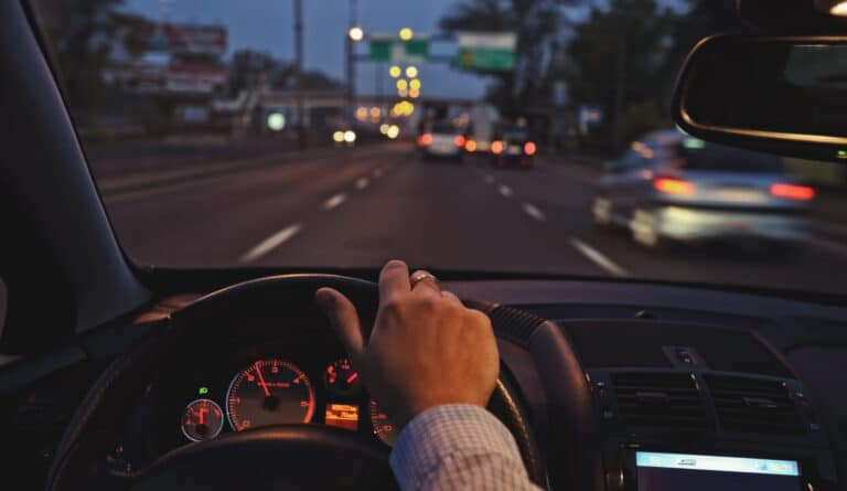 Driver grips steering wheel at night, dashboard lit, city traffic ahead with blurred lights. Driver grips steering wheel at night, dashboard lit, city traffic ahead with blurred lights.