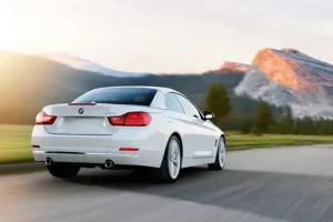 White BMW coupe driving on scenic mountain road at sunset with pines and clouds.