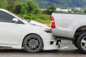 White car rear-ends gray pickup; vehicle damage, blurred greenery behind.