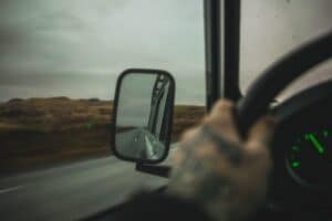 Tattooed hand on steering wheel, empty road in side mirror, cloudy sky outside.