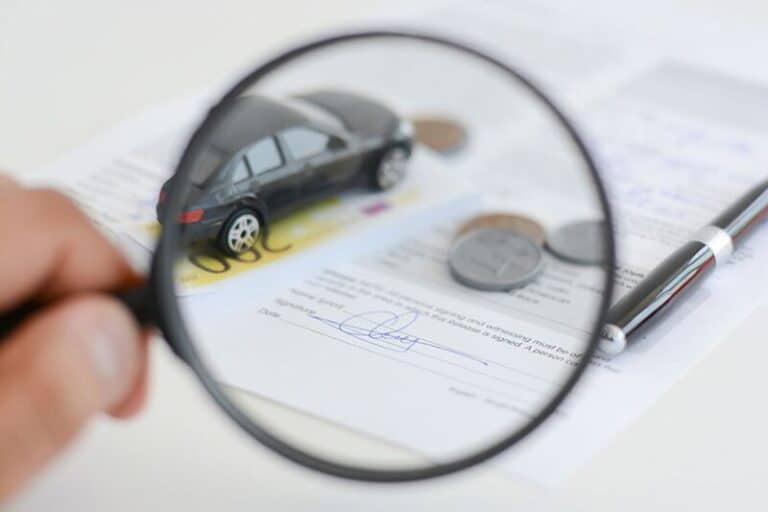 Magnifying glass over car insurance paper, with toy car, coins, pen, and euro note.