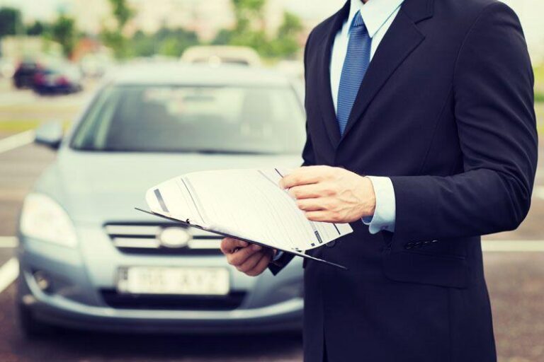 Man in suit with clipboard by gray parked car; face out of view.