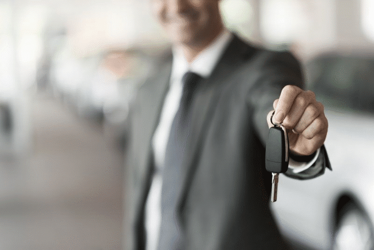 Smiling person in suit offers car keys in showroom; cars blurred behind.