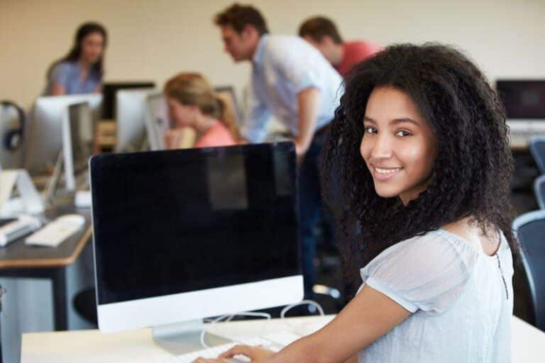 Smiling student at desk with computer; classmates work in background.