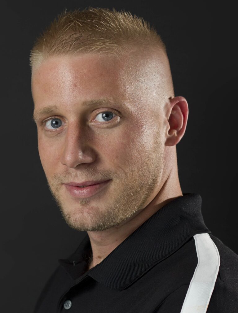 Man with short blond hair in black polo smiles at camera, dark backdrop.