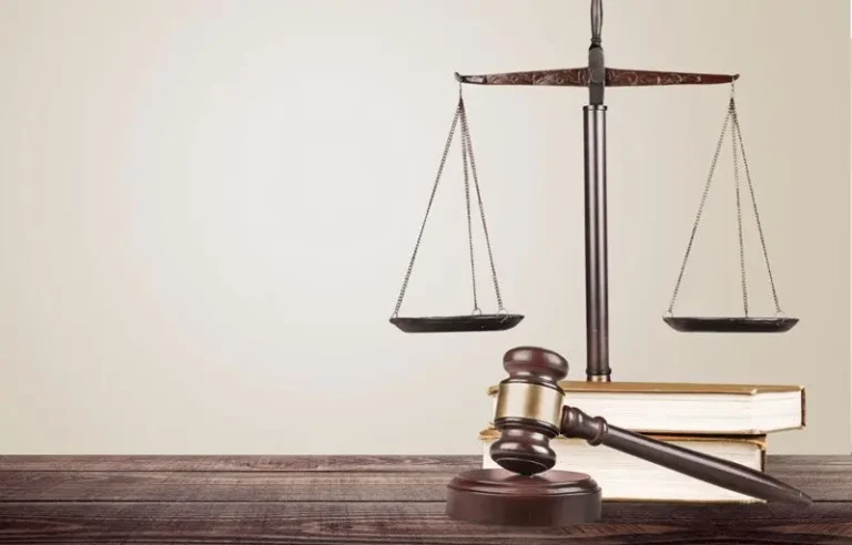 Gavel on block, books, and balance scale on wooden table against light wall.