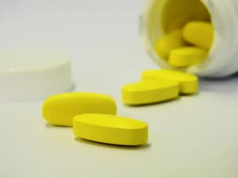 Yellow pills scattered near an open white pill bottle and cap on a white surface.