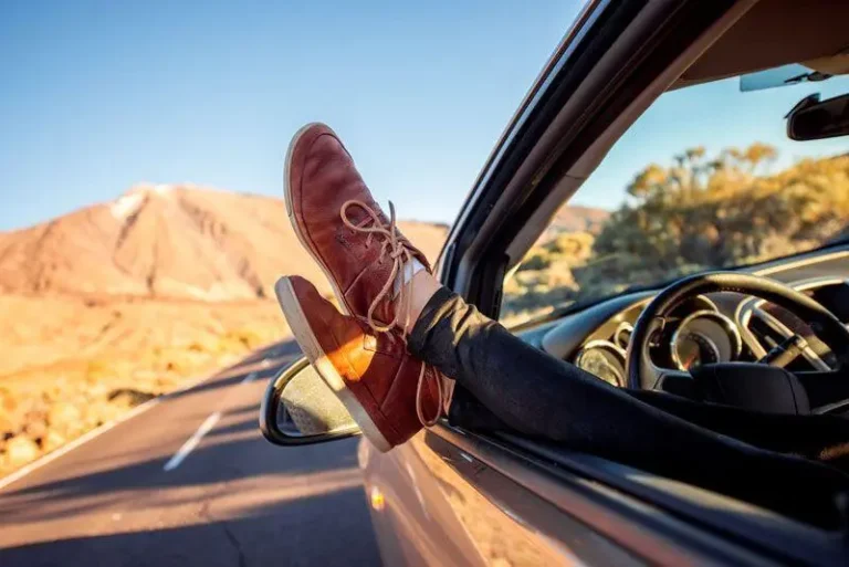 Legs with brown shoes out car window, mountains and blue sky in background.