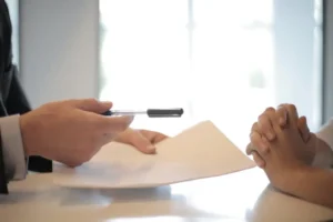 Person in suit gives pen and papers for signing across a bright office table.