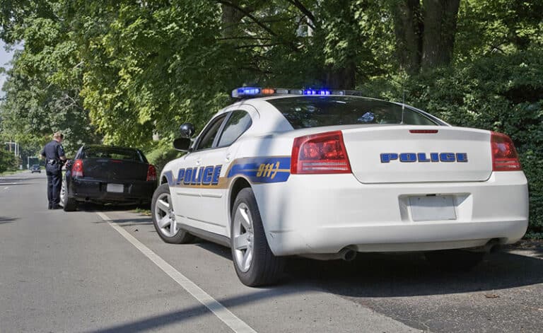 Police officer and black car pulled over, police lights flashing on a tree-lined road.