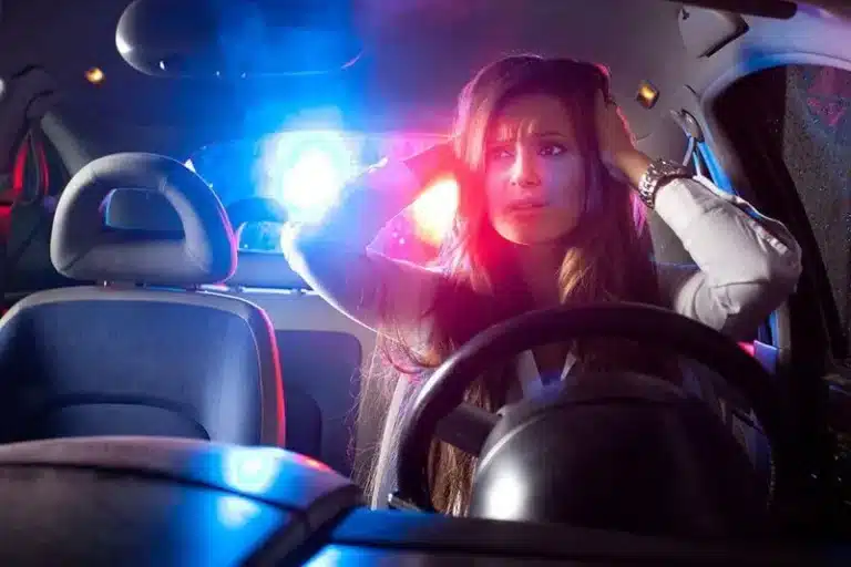 Woman in car at night, hands on head, worried; police lights flash behind.