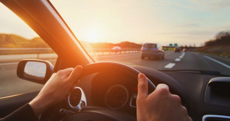 Driving on a sunset highway, hands on wheel, cars ahead, warm sun glow.