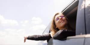 Person in sunglasses smiling, arm out car window, enjoying a sunny day.