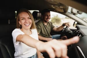 Smiling woman points out car window, man drives; both enjoy a road trip.