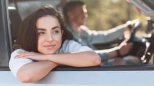 Smiling woman leans on car window; sunlit, blurry scenery outside.