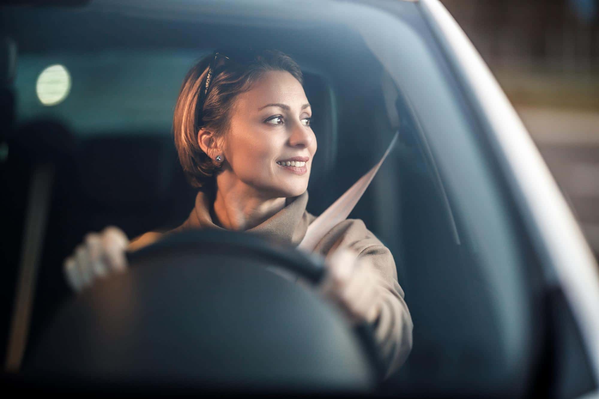 Woman in tan coat smiles, driving and looking out car window.