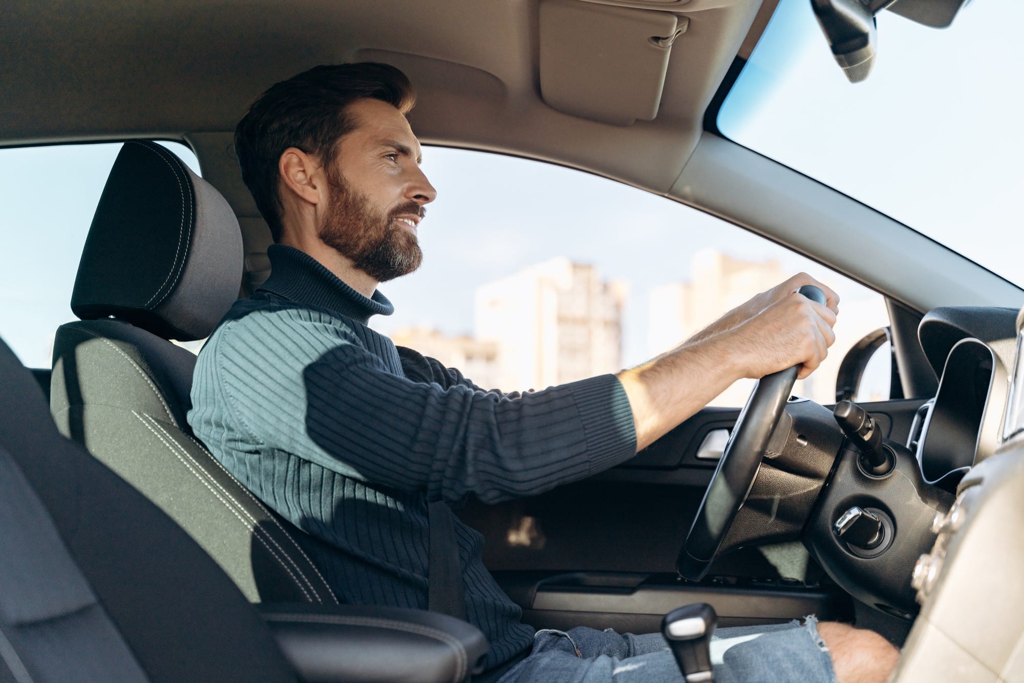 Bearded man driving car, looking ahead with city buildings outside window.