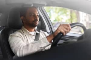 Man in light shirt driving, focused, with sunlight and green trees outside window.